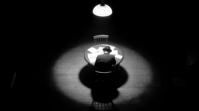 black and white film noir photograph from slightly above, single figure at a minimal desk working under a hanging pendan